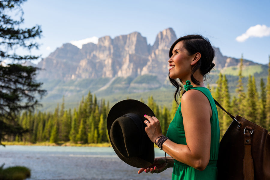 A traveler holding a black hat stands beside a lake with mountains behind her.
