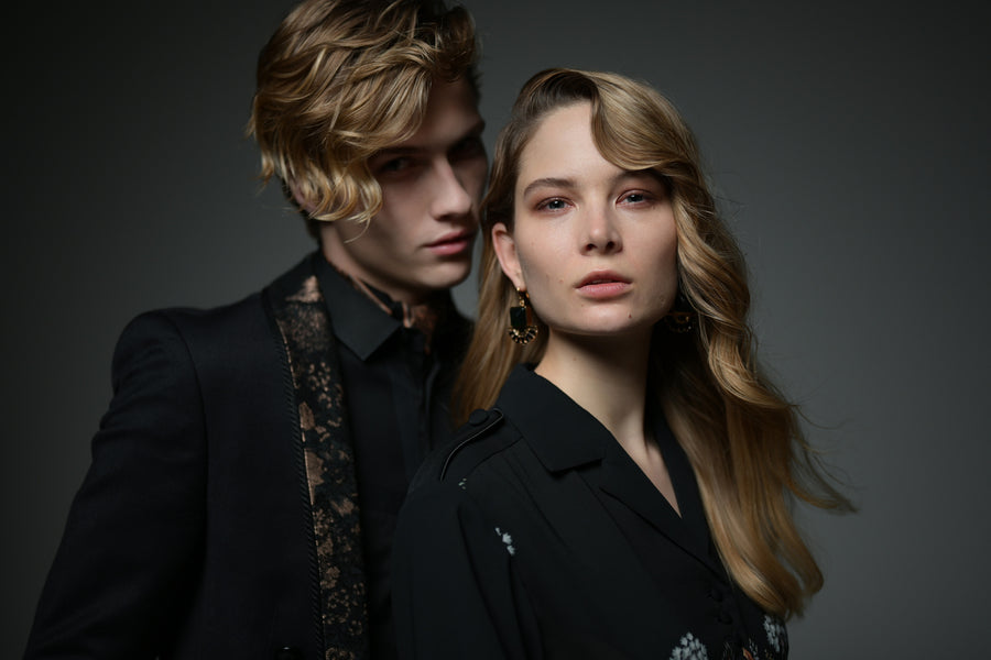 Two people in dark clothing pose together against a softly lit dark background.
