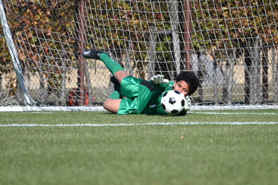 A goalkeeper stretches across the grass to stop a soccer ball near the goal.